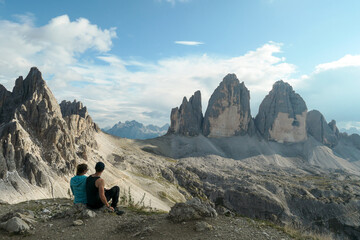 A couple in hiking outfit, sitting on the ground and enjoying the view on the famous Tre Cime di Lavaredo (Drei Zinnen) in Italian Dolomites. Desolated, raw landscape, full of lose stones. Sunny day