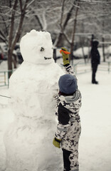 Lifestyle photo of a boy putting a carrot as a nose on snowman on snowy winter day outdoors....