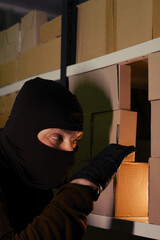 A man in a ski mask picks up a box with a parcel in a warehouse at night