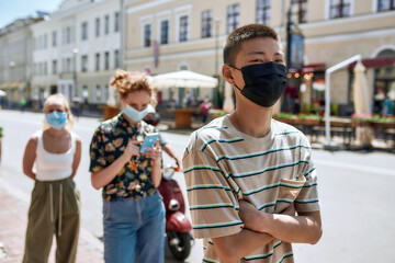 Young asian guy wearing mask waiting in line, respecting social distancing to enter takeout restaurant or to collect purchases from the pickup point during coronavirus lockdown