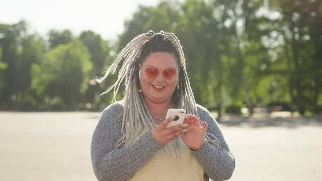 Smiling happy woman with dreads texting message standing at city street during summertime. Stout girl or mobile user looking at mobile screen surfing in internet. Lifestyle and social media concept