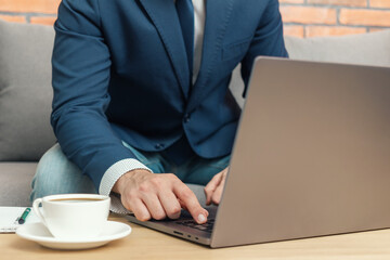 Unrecognized man, businessman or freelancer sitting on sofa and working at laptop on table with coffee cup, work from home