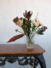 Bouquet of fresh flowers of beige brown green shades in a crystal vase on a wooden vintage table against a light wall background

