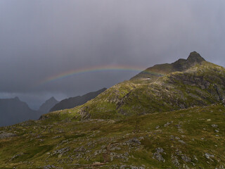 Panoramic view of Munkebu peak on Moskenesøy island, Lofoten, Norway, Scandinavia with green meadows, rugged mountains and beautiful colorful rainbow on stormy day in summer.
