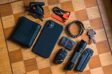 EDC setup. Concept on the wooden chess board. Everyday carry for a man. Knife, Multitool, Flashlight, Car key, Paracord bracelet, Wallet, Lighter, Smartphone...