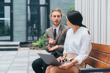 asian girl and man in business clothes on a bench with a laptop are discussing
