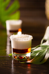Tropical alcoholic shots decorated with coconut flakes, monstera flower with coconut and pineapple on brown wooden background