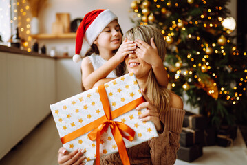 Fototapeta na wymiar A cute little girl is giving his handsome mother a gift box. Cheerful mom and her cute daughter girl exchanging presents. Parent and small child have fun near the tree indoors. Loving family.