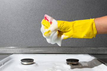 Woman's hand in glove cleaning gas stove. Woman squeezes foam from sponge. Kitchen cleaning, household chores