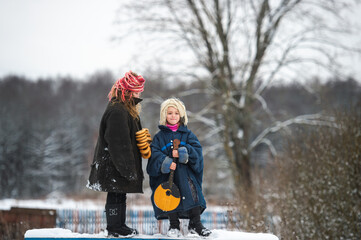 Two beautiful village girls stand on the bench with balalaika and barankas in winter