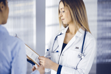 Young woman-doctor and her patient are discussing patient's current health examination, while standing together in a hospital office. Female physician is writing some marks, using a clipboard. Perfect