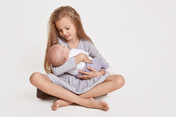 Blond female kid wearing grey dress sitting on floor with crossed legs sitting on floor and holding her infant sister in hands, looking at her with great love.