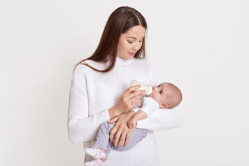 Mother feeding her baby from feeding bottle, dark haired female wearing sweater hold baby in hands and looking at her kid, posing isolated over white background.