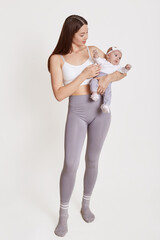 Full length portrait of mother standing and holding baby in hands, lady wearing bra and gray leggins, posing isolated over white background, mommy playing with infant girl.