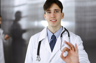 Friendly man-doctor standing straight and showing Ok sign in clinic. Female colleague is in a hurry at the background of physician. Medicine concept