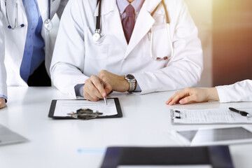 Fototapeta premium Group of unknown doctors are sitting at the desk and discussing problems or disease treatment in sunny hospital office. Covid 2019