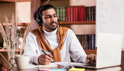 Serious Indian Teacher Sitting At Laptop Wearing Headphones Indoors