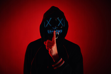 Anonymous man in hoodie hiding face behind neon glow scary mask on red background. Horror concept