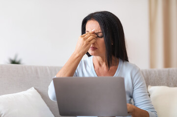 Fatigued upset mature woman massaging nose bridge, using laptop
