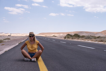Blond woman sits on the road highway looks away on road,  in hat and yellow t-shirt. time of travel, space for text. road in desert. no have cars.