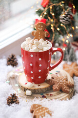 A cup of hot chocolate toped with marshmallow, gingerbread man biscuit at a window. With  Christmas sugar cookies decorated with royal icing and xmas tree light decoration atmosphere in background  