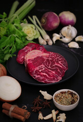 A part of the raw red beef shank in a black plate with ingredients such vegetables (lemon grass, celery, garlic, onion),herbs and spices to preparing for cooking a traditional Asian broth soup      