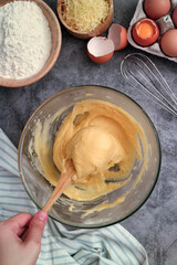 Top view of uncooked French chou pastry dough and hands batting with paddle in a mixing bowl. Preparation step of traditional French gougères with ingredients such flour ,butter ,grate cheese, eggs. 