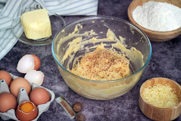 An uncooked French chou pastry dough with grated cheese, grounded nutmeg in a mixing bowl. Ingredients as butter, eggs, flour in scene. Step of preparation. A traditional pastry made with savory choux