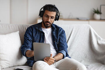 Indian guy spending time at home with digital tablet and wireless headphones - obrazy, fototapety, plakaty