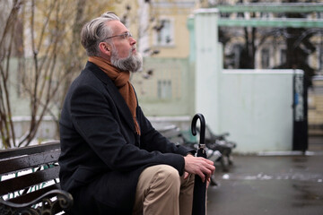 A respectable adult man with a gray beard sits on a park bench and enjoys the weather