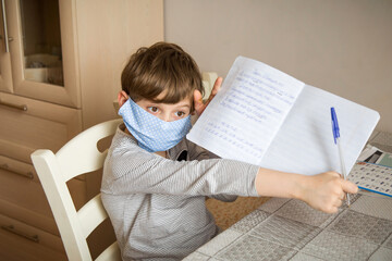 A first-grader boy in a mask is doing homework in quarantine because of a coronavirus.