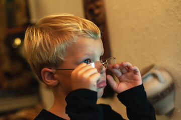 A bespectacled preschool boy straightens his glasses on his nose with two hands. Looks over glasses