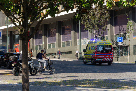 Porto, Portugal - 08/28/2019: Red and yellow ambulance responding to an emergency. Men stopped in their bikes and people walking in the sidewalk.