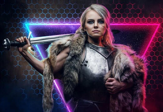 Powerful and beautiful female warrior with gray hairs and scar in steel vest with fur poses in abstract background holding a sword on her shoulder..