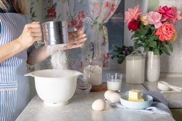 housewife in an apron sifting flour into bowl. preparing dough for home baking. cooking at home, hobbies. close-up.