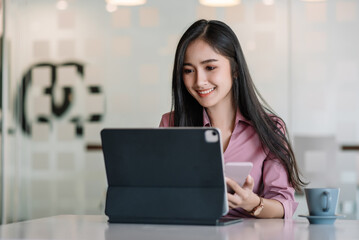 Young asian businesswoman beautiful charming smiling and using tablet and mobile phone, while sitting at table and working in office.