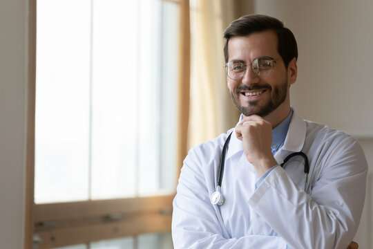 Profile picture of smiling young Caucasian male GP or pediatrician in white medical uniform and glasses pose in private clinic. Portrait of happy positive man doctor in eyewear. Medicine concept.