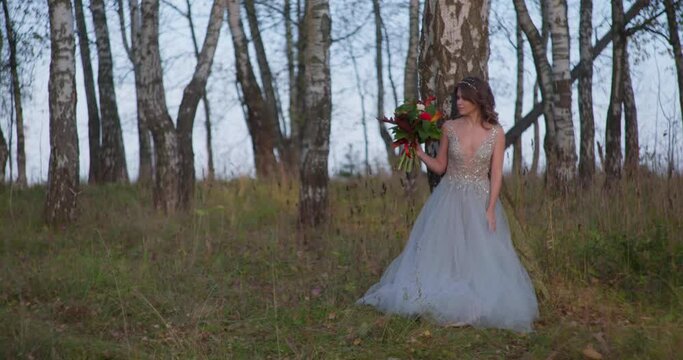 A bride in a gray dress walks with a wedding bouquet in the forest.