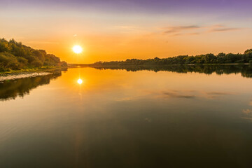 Scenic view at beautiful summer river sunset with reflection on water with trees , golden sun rays, calm water ,deep blue cloudy sky and glow on a background, spring evening landscape