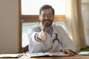 Fototapeta premium Close up blurred of smiling male doctor stretch hand greeting with patient or client at consultation in hospital. Man GP sit at desk get acquainted meet customer in private clinic. Medicine concept.