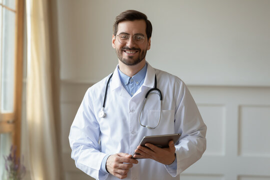 Profile picture of smiling young Caucasian male doctor in white medical uniform and glasses in hospital. Headshot portrait of happy man GP use tablet in private clinic. Healthcare, medicine concept.