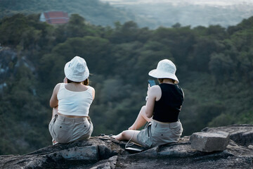 Two beautiful Asian girls sit on a cliff overlooking nature and mountains.