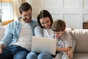 Overjoyed young parents with small son relax on sofa in living room laugh watching funny video on laptop. Smiling family with little boy child enjoy weekend at home, have fun use modern computer