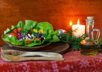 Christmas still life with salad of tomato, cheese, green onions and tricolor violet flowers,  a candle,  wooden spoons, a branch of juniper and a bottle of olive oil