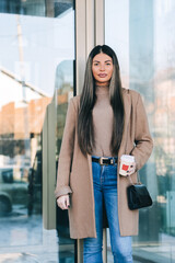 Beautiful young stylish woman wearing beige coat holding paper coffee cup and enjoying the walk in the city. Autumn, winter luxury look