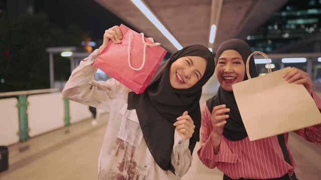 Two young Asian muslim friends feel happy  showing shopping bags to camera, Modern Muslim lifestyle, capitalist consumer spending money, discounts sales products, enjoy shopping at the mall, city life