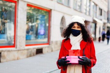 Portrait of a woman in red shopping outside on the street with face mask walking and holding many christmas gifts and presents looking at camera