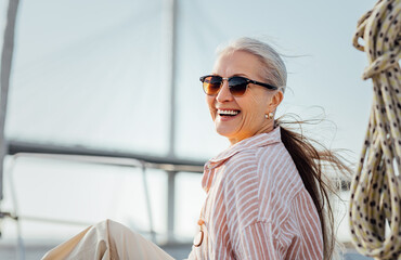 Laughing mature woman wearing sunglasses and looking at camera. Smiling female in casuals enjoying vacation. - obrazy, fototapety, plakaty