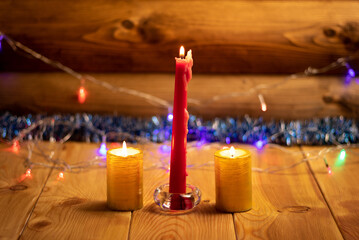 One long red candle stands between two Golden ones in the center of the photo. Candles are placed on a wooden table. In the background-a wall of logs and a Christmas garland. Perfect Christmas mood.