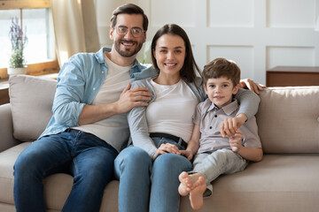 Portrait of happy young Caucasian family with little preschooler son sit relax on sofa in living room. Smiling parents mom and dad rest on couch at home, enjoy weekend together with small boy child.